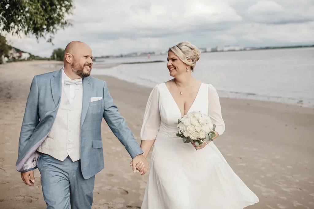 Brautpaar spaziert Hand in Hand am Elbstrand in Hamburg – Hochzeitsfotos mit maritimem Flair Das Brautpaar Christina und Andre genießt einen emotionalen Moment am Elbstrand in Hamburg. Der Bräutigam im hellblauen Anzug und die Braut mit weißem Kleid und Rosenstrauß lächeln sich an, während sie barfuß am Wasser entlanggehen. Diese Hochzeitsaufnahme fängt die Leichtigkeit und Romantik einer freien Trauung im Norden perfekt ein – mit der Elbe im Hintergrund und dem typischen Hamburger Himmel darüber.